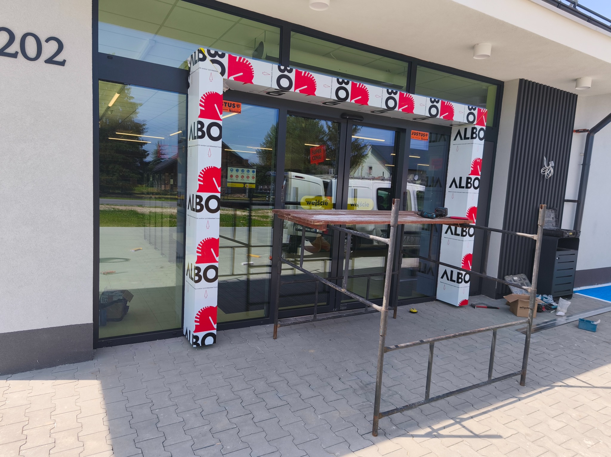 Montaż obudowy wejścia do sklepu z płyt kompozytowych ALBO. Widoczne rusztowanie i narzędzia. Prace w toku, elewacja budynku z elementami dekoracyjnymi.