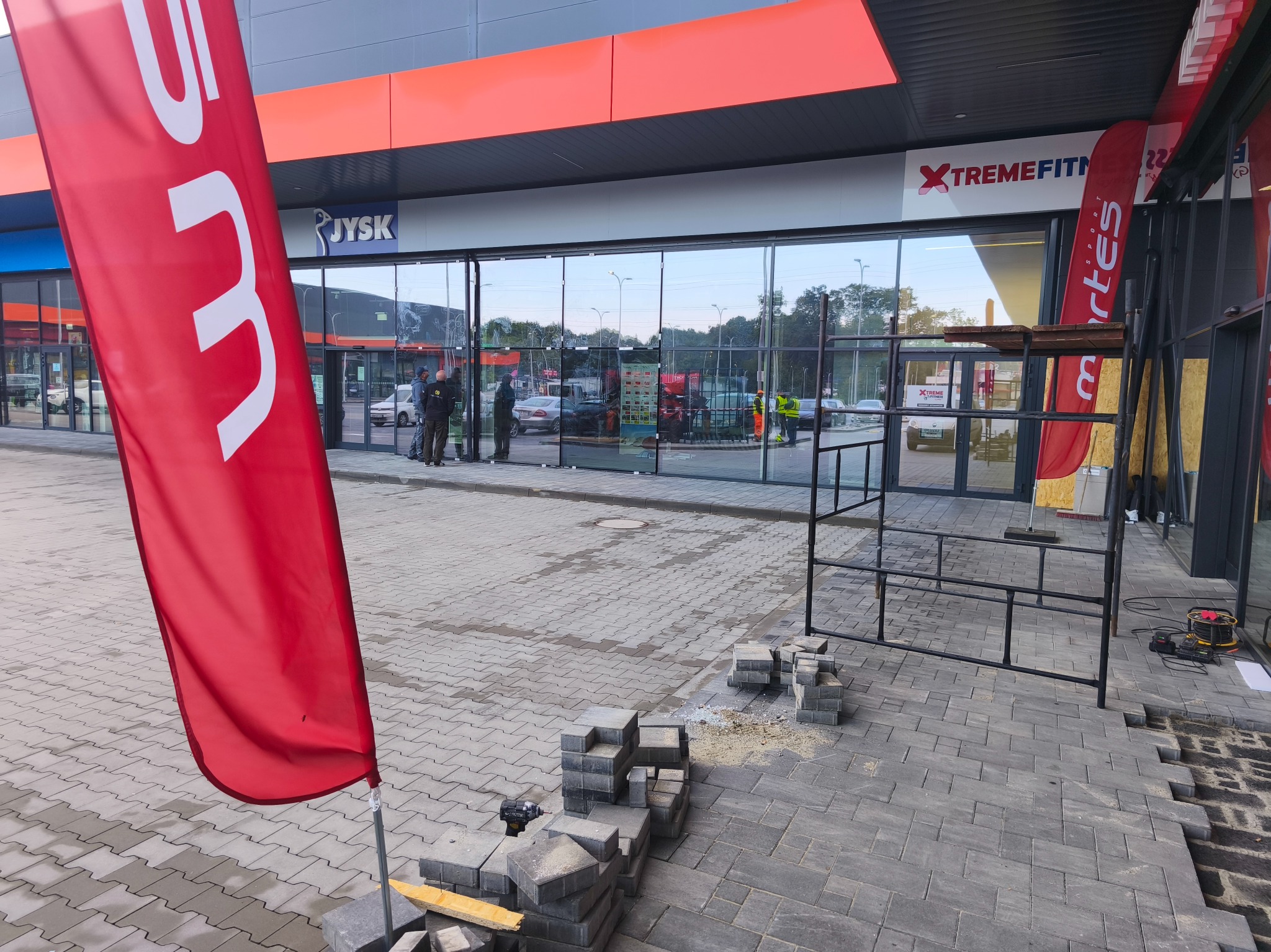 Układanie szarej kostki brukowej przed budynkiem JYSK i XTREMEFITNESS. Widoczne narzędzia i częściowo ułożona nawierzchnia. Prace brukarskie w toku.