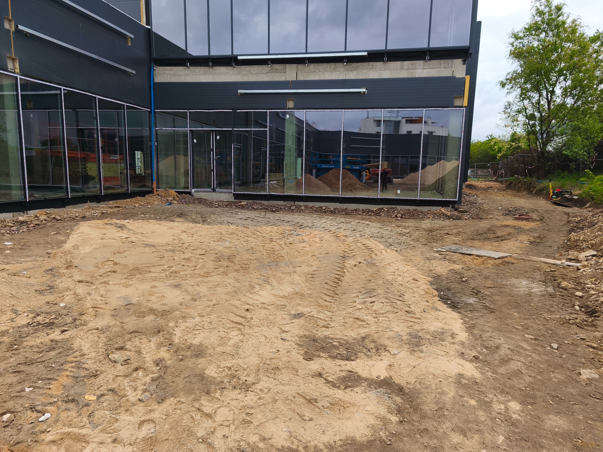 Budynek komercyjny z szarą elewacją i dużymi oknami, w trakcie prac ziemnych. Widoczne hałdy piasku i sprzęt budowlany w odbiciu okien.