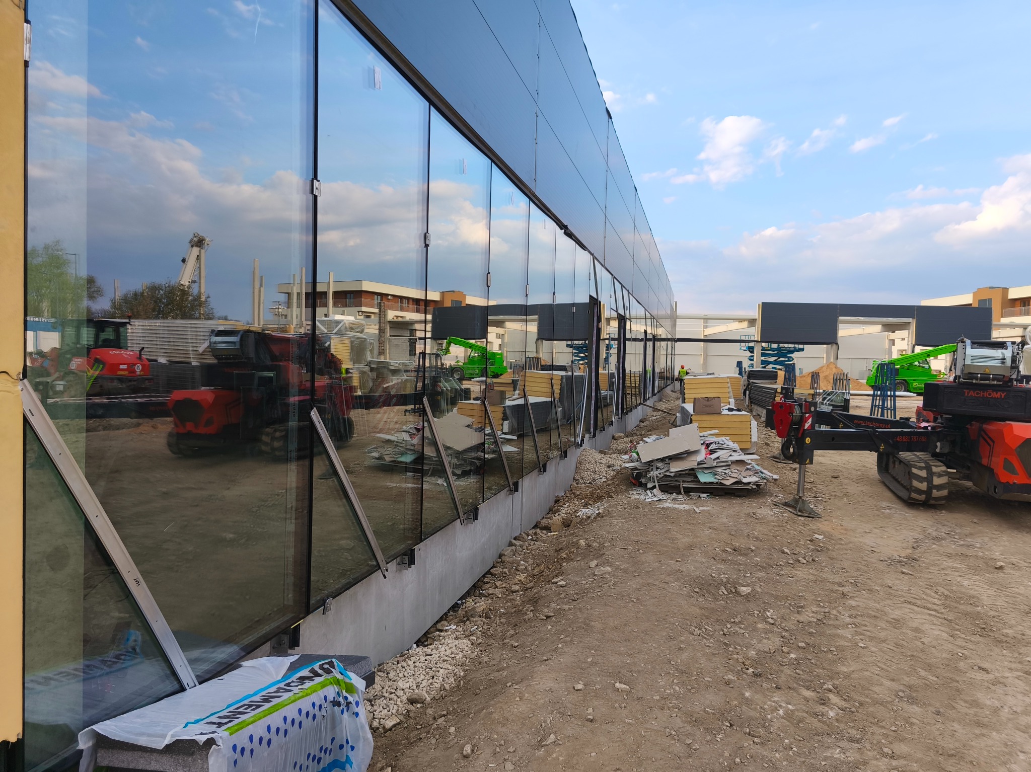 Montaż szklanej fasady budynku w trakcie realizacji. Widoczne maszyny budowlane i materiały. W tle odbicie nieba i budynków w szklanej powierzchni.