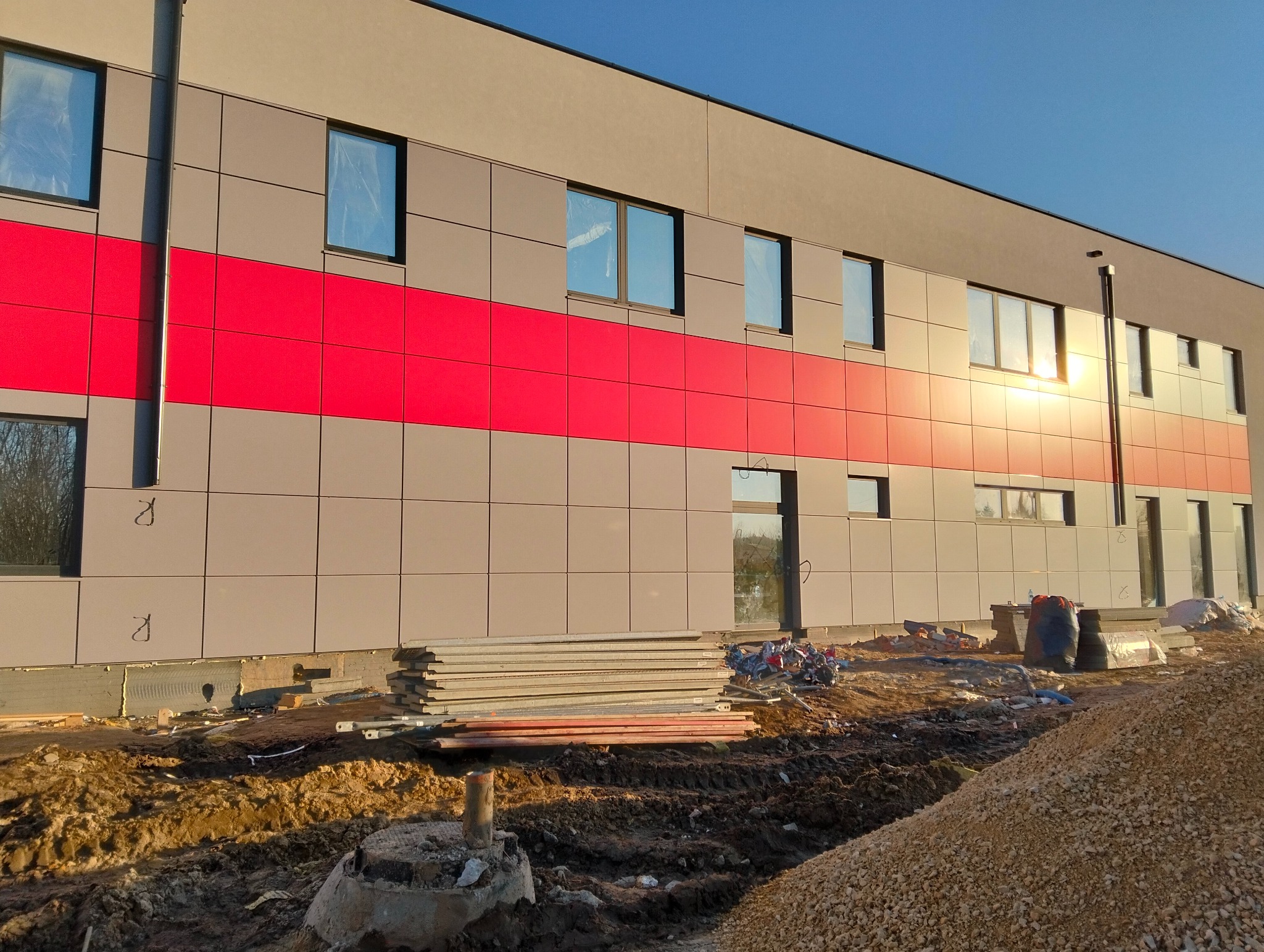 Elewacja budynku z panelami w Bielsko-Białej, z wyraźnym czerwonym pasem. Widoczne okna i stos paneli na ziemi, sugerujące prace wykończeniowe.