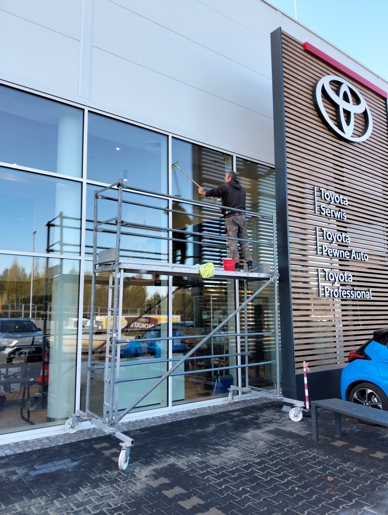 Pracownik myje okna na rusztowaniu przed salonem Toyoty. Widoczne logo i napisy informacyjne. Jasne, duże okna i czyste otoczenie.
