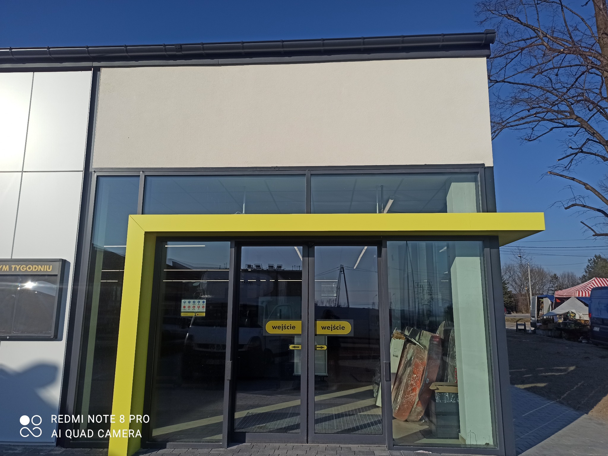 Nowoczesny budynek z przeszklonym wejściem, żółtą ramą i szarymi panelami elewacyjnymi. Widoczne drzwi z napisem 'wejście' i fragment drzewa na tle błękitnego nieba.