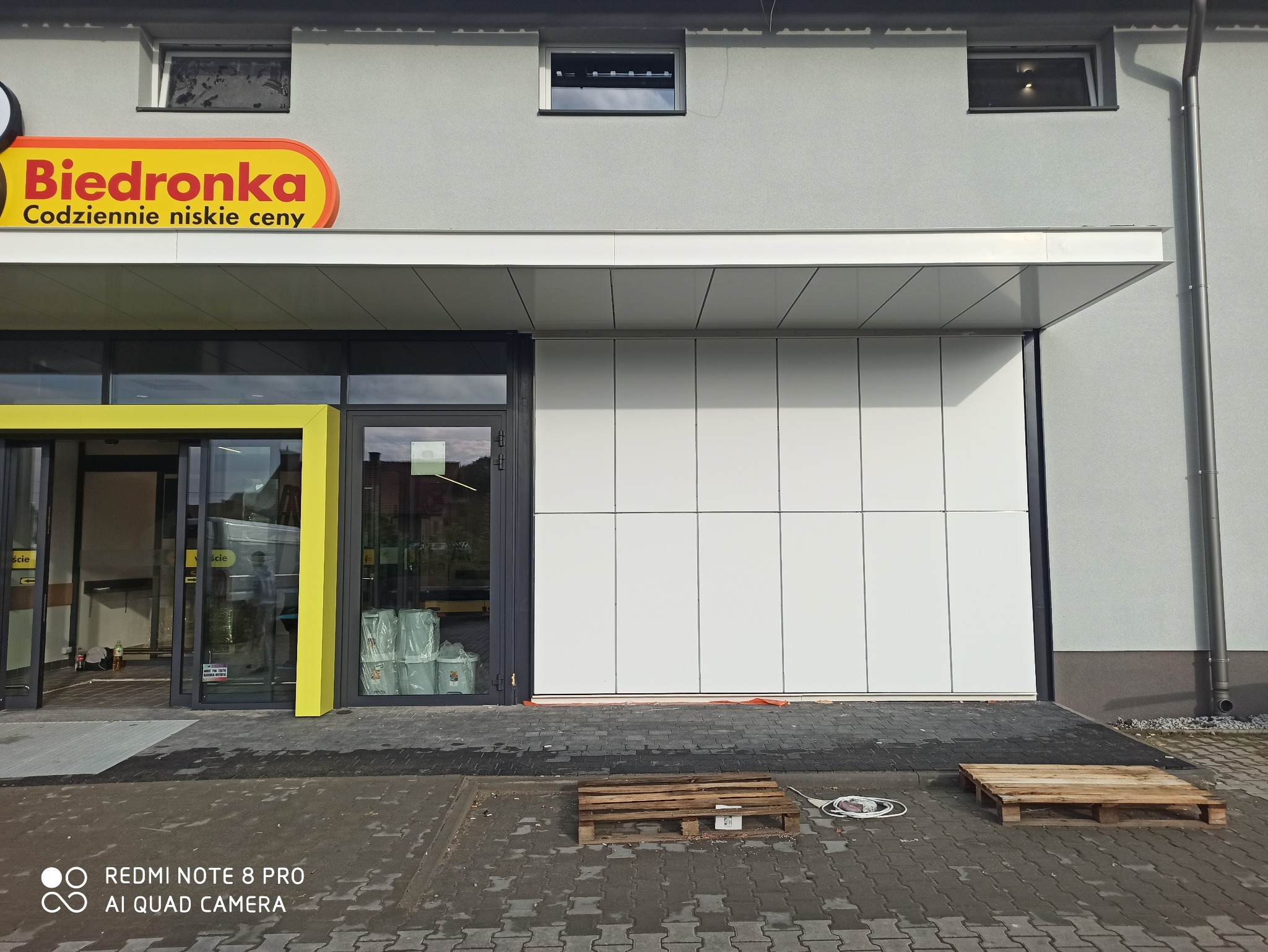 Elewacja budynku sklepu z zamontowanymi panelami elewacyjnymi. Widoczne wejście z żółtą ramą i palety na chodniku. Ujęcie z poziomu chodnika.