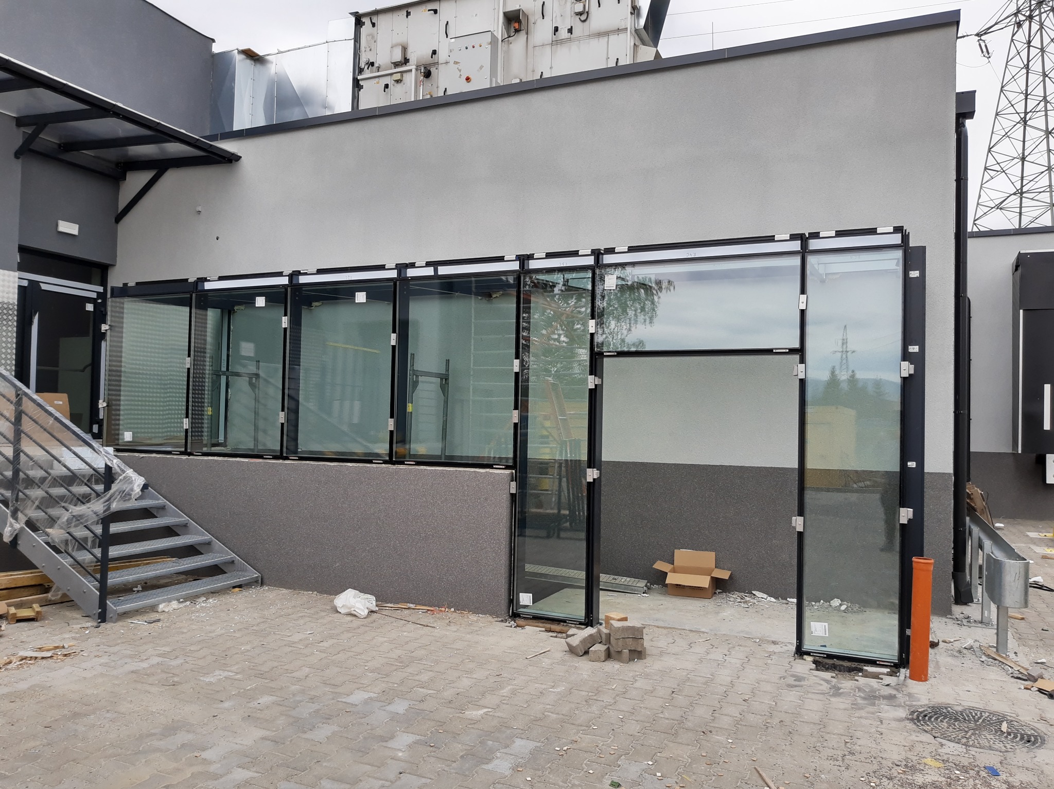 Szklana fasada budynku w trakcie montażu, widoczne elementy konstrukcyjne i otwarte przestrzenie. Na pierwszym planie kostka brukowa i schody.