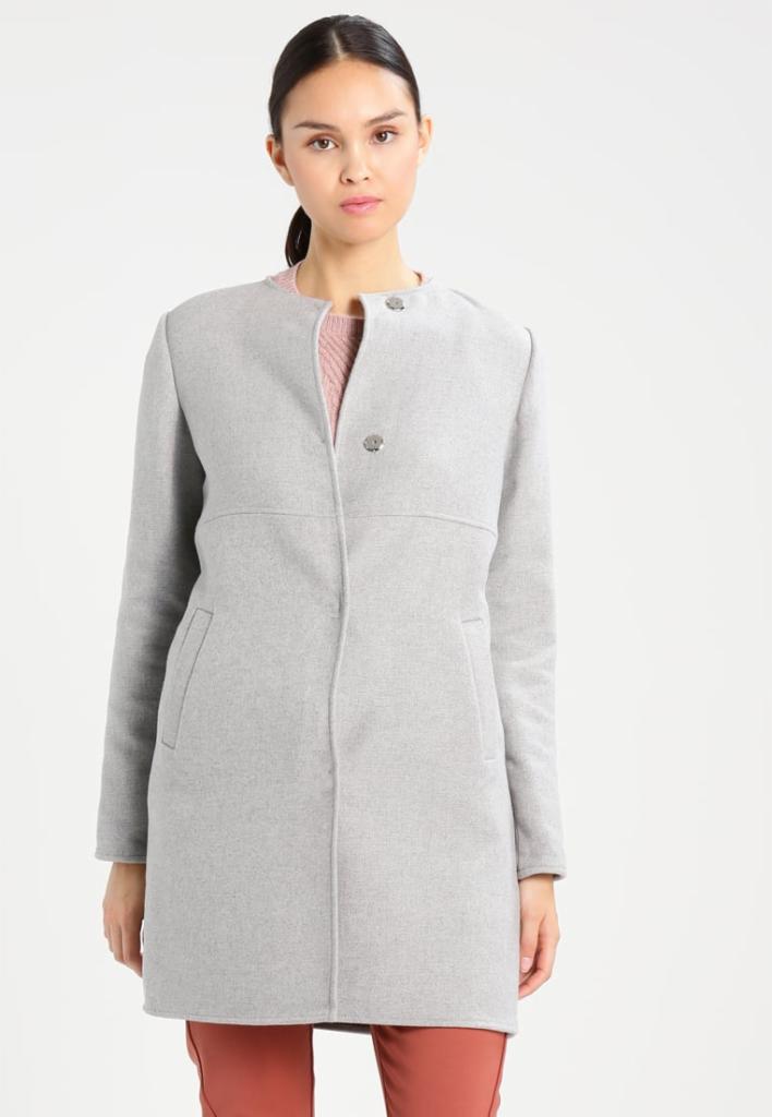 Kobieta prezentuje szary, wełniany płaszcz z okrągłym dekoltem i ozdobnymi guzikami, widoczny fragment różowego swetra pod spodem.