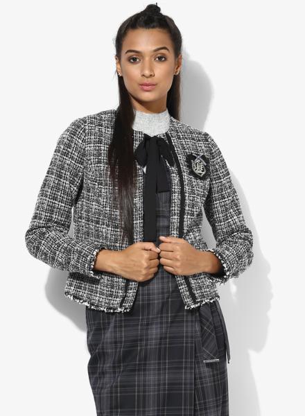 Kobieta w tweedowej marynarce bez zapięcia z ozdobną naszywką i czarną wstążką, ubrana w szarą sukienkę w kratę i golf.