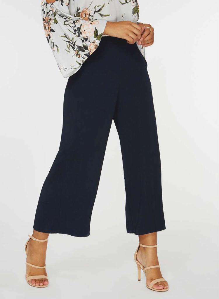 Eleganckie, granatowe spodnie typu culottes z wysokim stanem, zestawione z kwiecistą bluzką z bufiastymi rękawami i beżowymi sandałami na szpilce.