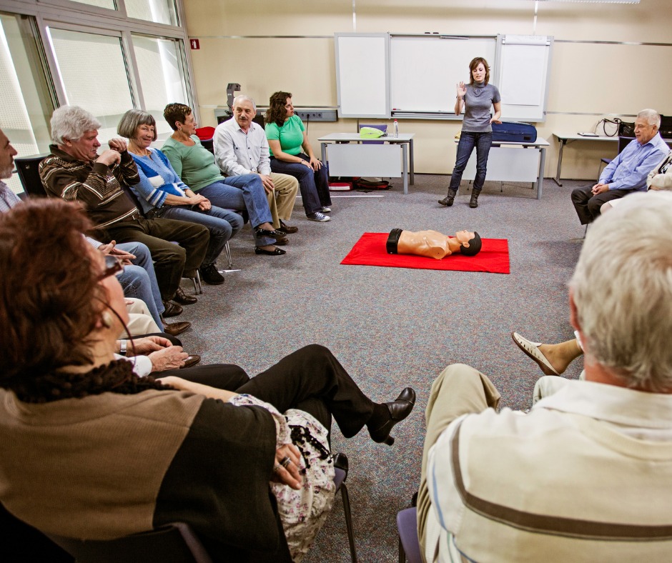 Grupa osób siedzi w kręgu podczas szkolenia, z manekinem na czerwonym kocu pośrodku, instruktor gestykuluje przy tablicy.