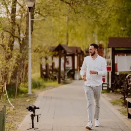 Kamerzysta w jasnej koszuli i kraciastych spodniach idzie ścieżką w parku obok kamery na statywie.