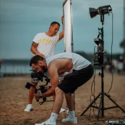 Ekipa filmowa na plaży: operator z kamerą na stabilizatorze, asystent z blendą i lampą, w tle woda i zachmurzone niebo.