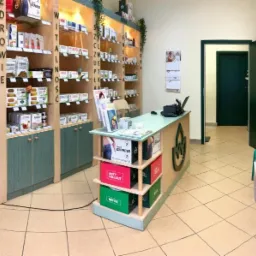 Wnętrze gabinetu z regałami pełnymi produkt&oacute;w zdrowotnych, zielonymi akcentami kolorystycznymi, ladą z logo i poczekalnią z krzesłami.