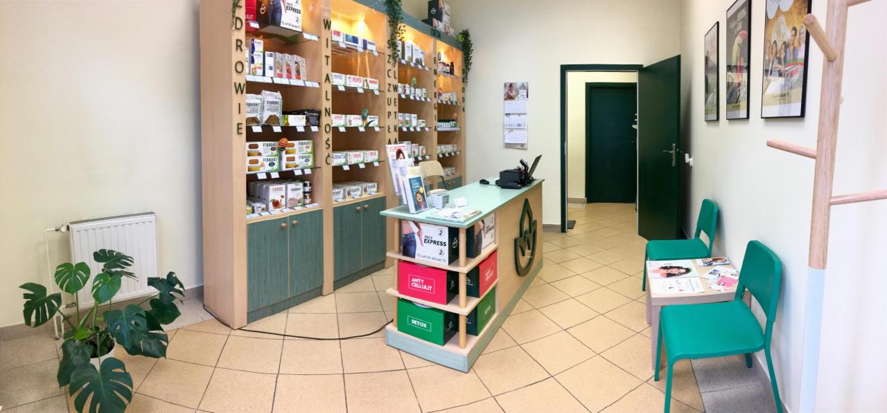Wnętrze gabinetu z regałami pełnymi produktów zdrowotnych, zielonymi akcentami kolorystycznymi, ladą z logo i poczekalnią z krzesłami.