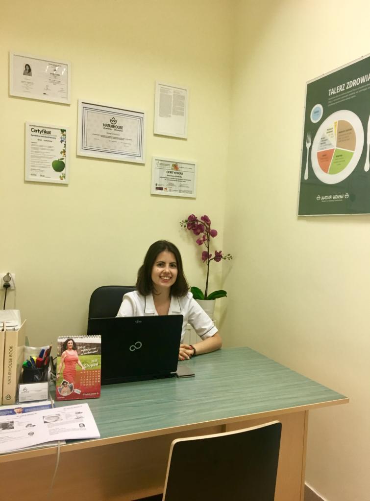 Uśmiechnięta kobieta w białym uniformie siedzi za biurkiem z laptopem w gabinecie, na ścianie wiszą certyfikaty i plakat 'Talerz zdrowia', obok storczyk w doniczce.