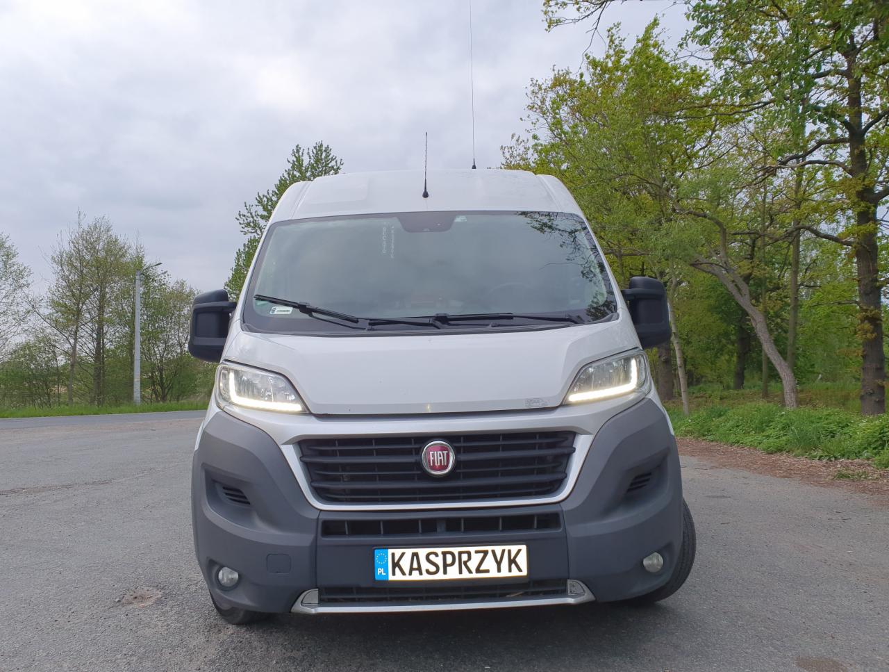 Przód białego busa Fiat Ducato z polską tablicą rejestracyjną KASPRZYK, stojącego na asfalcie w otoczeniu zieleni drzew.