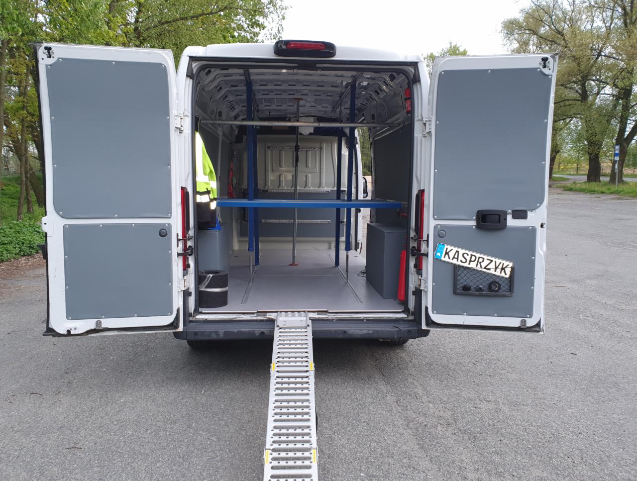 Wnętrze białego busa z otwartymi tylnymi drzwiami, wyposażonego w rampę wjazdową i specjalistyczne mocowania do transportu, widok od tyłu na szary asfalt.