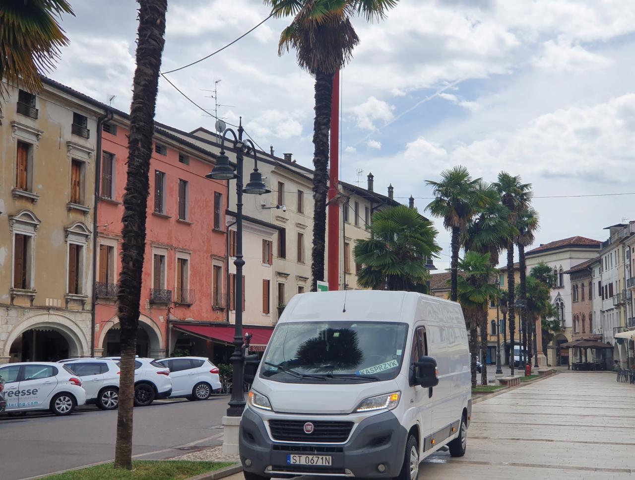 Biały bus dostawczy Fiat Ducato na wybrukowanym placu miejskim z palmami i kolorowymi kamienicami w tle, tablica rejestracyjna ST 0671N, naklejka z imieniem kierowcy na szybie.