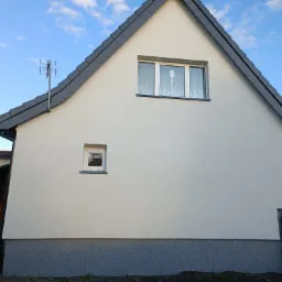 Świeżo otynkowana, biała elewacja domu jednorodzinnego z szarą podmurówką i dachem, widoczna antena telewizyjna.