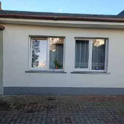 Świeżo otynkowana, pomalowana elewacja domu jednorodzinnego z dwoma oknami, na jednym z parapetów stoi butelka wody i doniczka z kwiatem, widoczna kostka brukowa przed budynkiem.