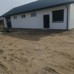 Biały, parterowy dom z ciemną dachówką i czarnymi roletami zewnętrznymi, widok z zewnątrz na niezagospodarowany teren z piaskiem i śladami opon.