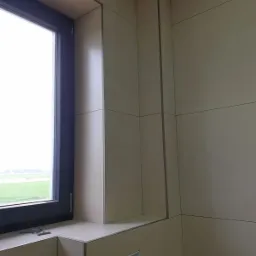 Beżowe płytki ścienne z ciemnymi fugami w łazience, widok na okno z czarną ramą i zielonym krajobrazem za nim, otwór instalacyjny w ścianie.