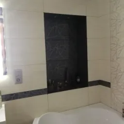 Wykończona łazienka z wanną, beżowymi płytkami z delikatnym wzorem liści i wnęką z ciemnymi płytkami, na której stoją kosmetyki i akcesoria.