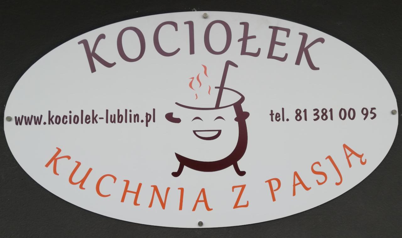 Owalny szyld reklamowy z nazwą 'Kociołek Kuchnia z Pasją', adresem strony internetowej, numerem telefonu i rysunkiem uśmiechniętego garnka z parującą zawartością.