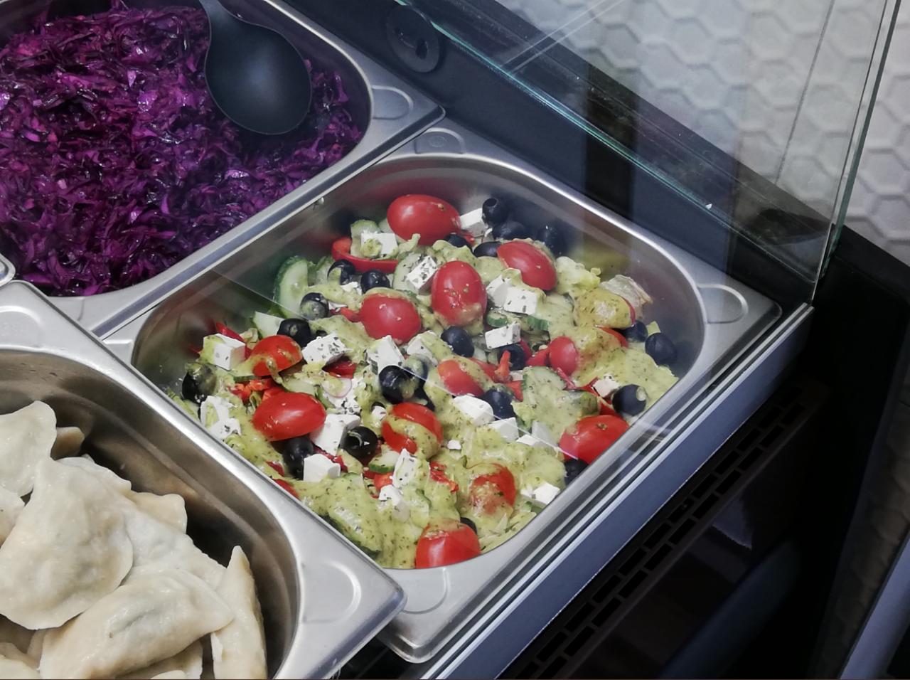 Metalowe pojemniki gastronomiczne z sałatką grecką, czerwoną kapustą i pierogami w witrynie chłodniczej.