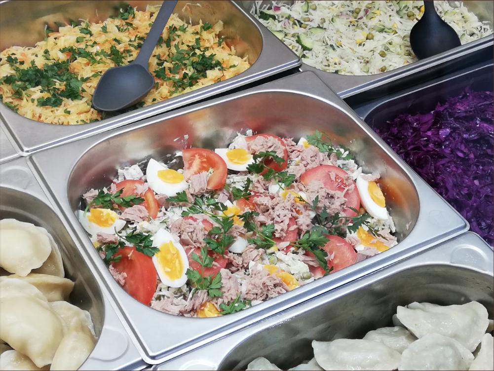 Metalowe pojemniki gastronomiczne z różnorodnymi sałatkami: jarzynowa z czarną łyżką, sałatka z tuńczykiem, pomidorami i jajkiem, sałatka z kapusty pekińskiej i zielonego ogórka, gotowana czerwona...