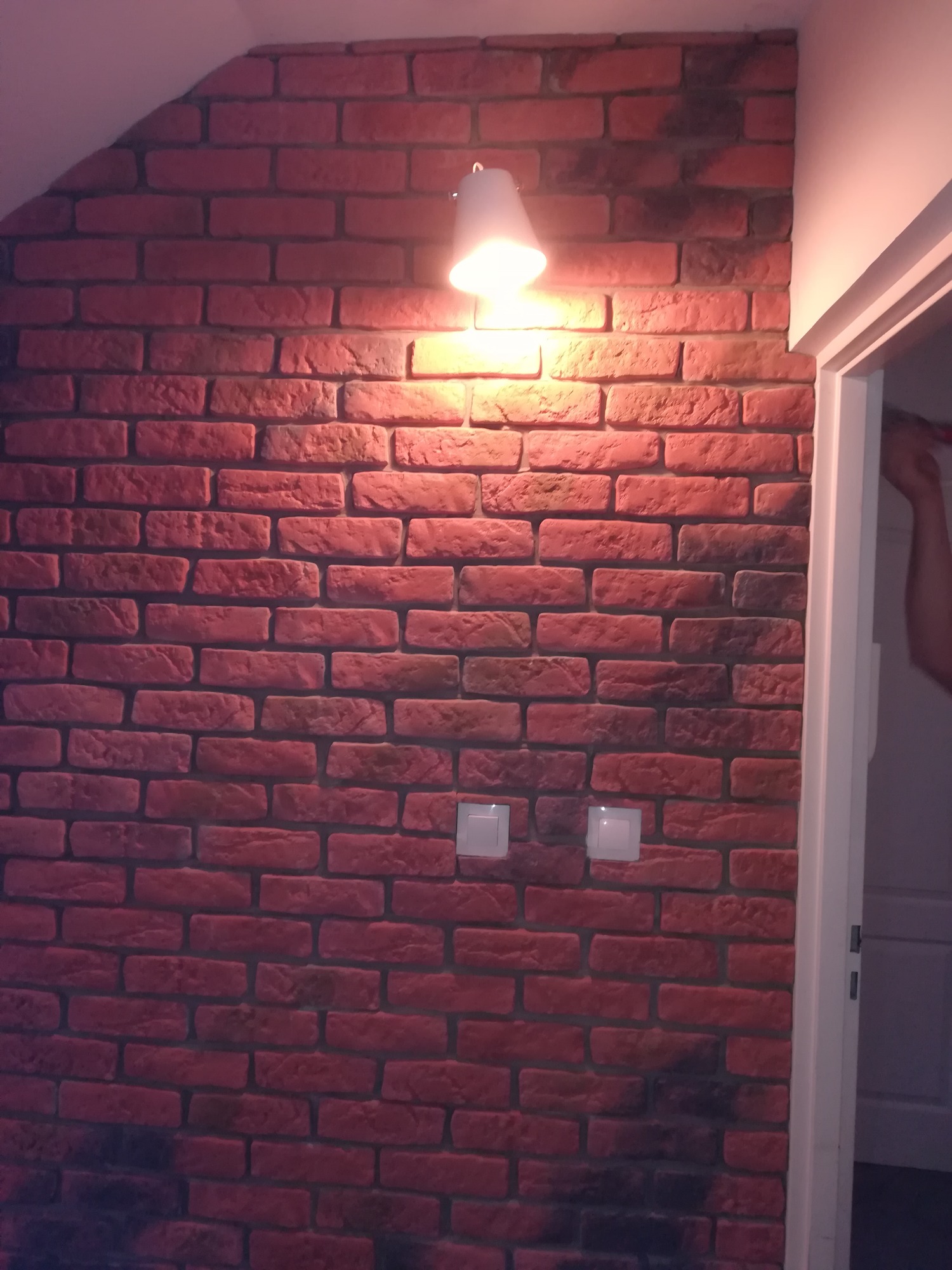Ściana pokryta czerwoną cegłą dekoracyjną z widocznymi dwoma włącznikami światła i lampą sufitową, oświetlona ciepłym światłem, z fragmentem białych futryn drzwi po prawej stronie.