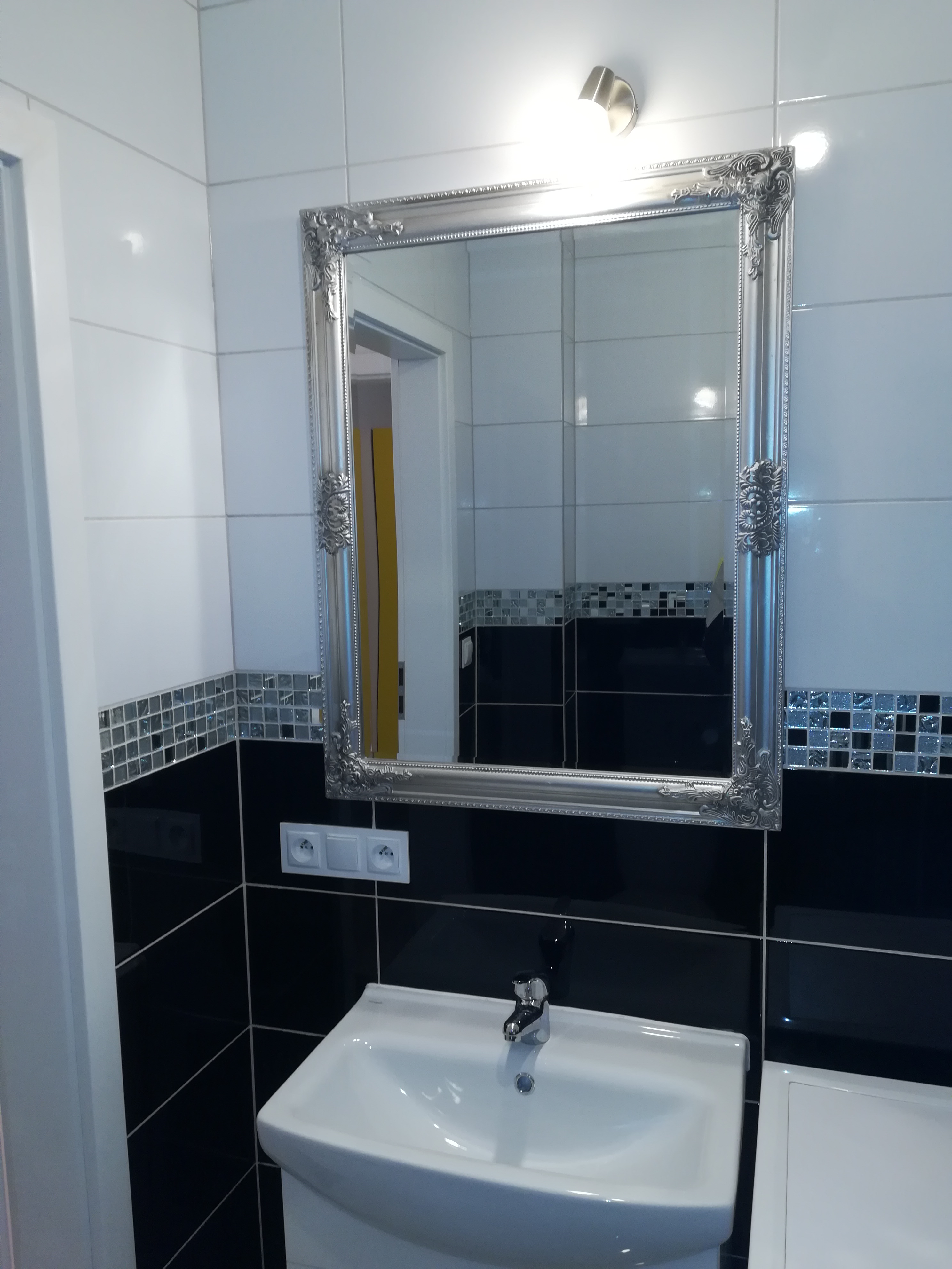 Wykończona łazienka z czarno-białymi płytkami, ozdobnym lustrem w srebrnej ramie, umywalką i baterią.