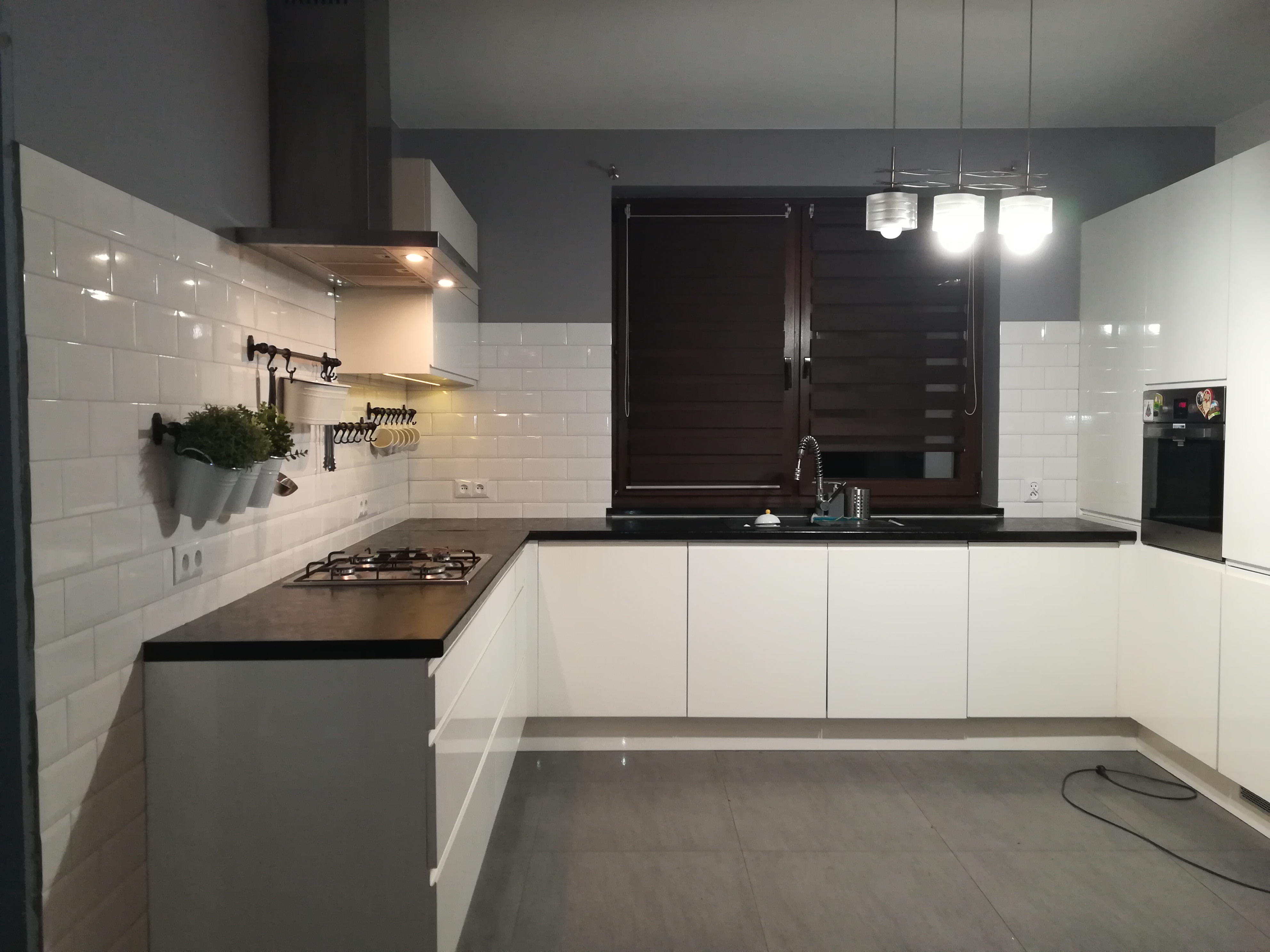 Nowoczesna kuchnia z białymi szafkami, czarnym blatem i białymi płytkami na ścianie, z wiszącymi doniczkami z ziołami i oświetleniem punktowym pod szafkami.