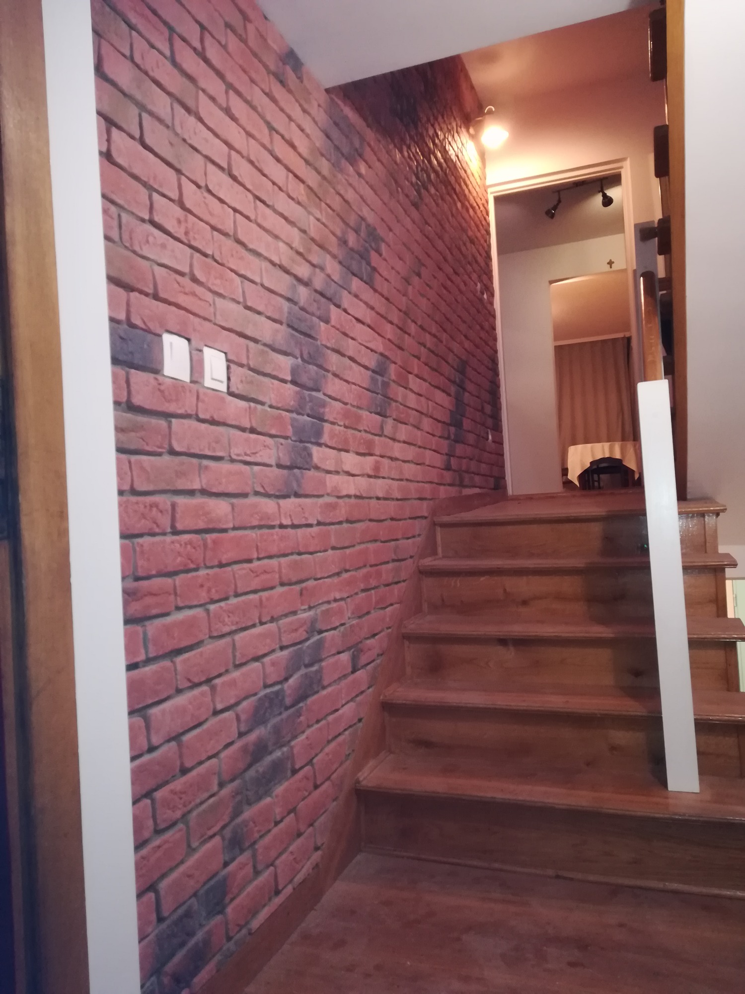 Schody drewniane prowadzące do pomieszczenia z widoczną ścianą wykończoną tapetą imitującą czerwoną cegłę, z widocznymi włącznikami światła.