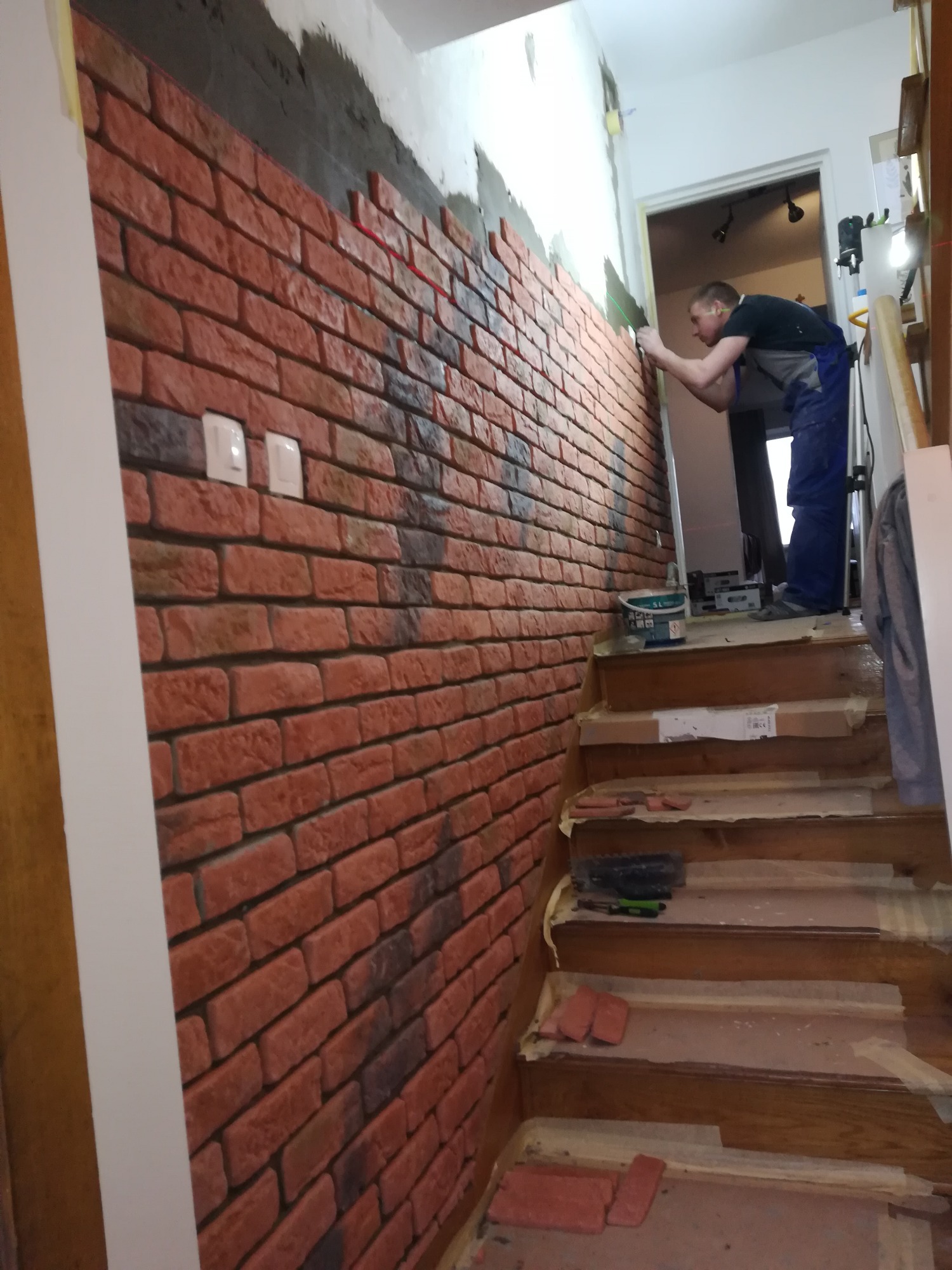 Montaż imitacji cegły na ścianie w korytarzu przy schodach. Widoczny pracownik w kombinezonie, używający poziomicy laserowej, oraz częściowo ułożona okładzina z cegły w odcieniach czerwieni i brązu...