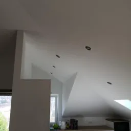 Pomalowane na biało poddasze ze skośnym sufitem, oświetleniem punktowym i zakrytą folią kuchnią w trakcie remontu.