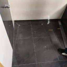 Ciemne, kwadratowe płytki podłogowe w kabinie prysznicowej z odpływem liniowym, białe płytki ścienne, widoczny fragment natrysku i czyjeś buty.