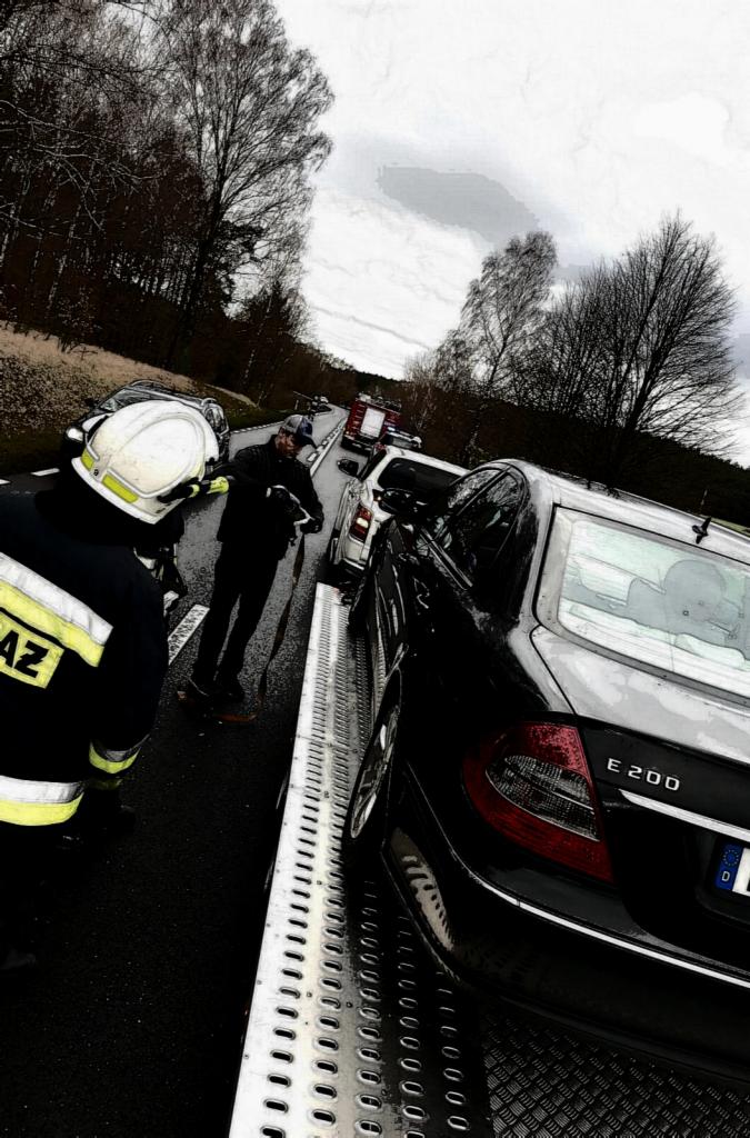 Czarny Mercedes E 200 na metalowej platformie lawety, strażak w białym hełmie i umundurowaniu z żółtymi pasami oraz mężczyzna w czarnej kurtce obserwują, w tle widać wóz strażacki.
