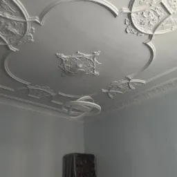 Ozdobny sufit z gładzią, sztukaterią i nowoczesną lampą wiszącą, widoczne zabezpieczone meble w tle.