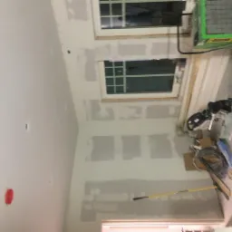 Ściana wewnętrzna z widocznymi łączeniami płyt kartonowo-gipsowych przed szpachlowaniem, narzędzia pracy i zielony osuszacz powietrza w tle.