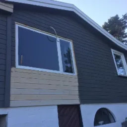 Dom w trakcie malowania elewacji na kolor grafitowy, z widocznym fragmentem surowego drewna i białym wykończeniem okien.