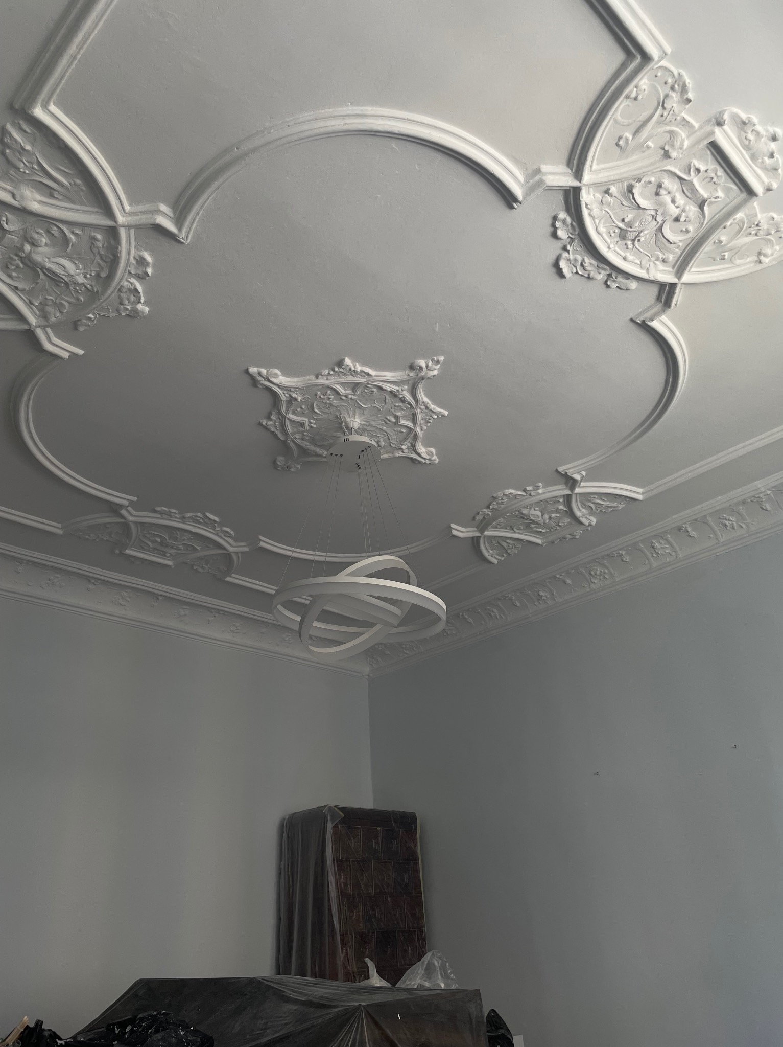 Ozdobny sufit z gładzią, sztukaterią i nowoczesną lampą wiszącą, widoczne zabezpieczone meble w tle.