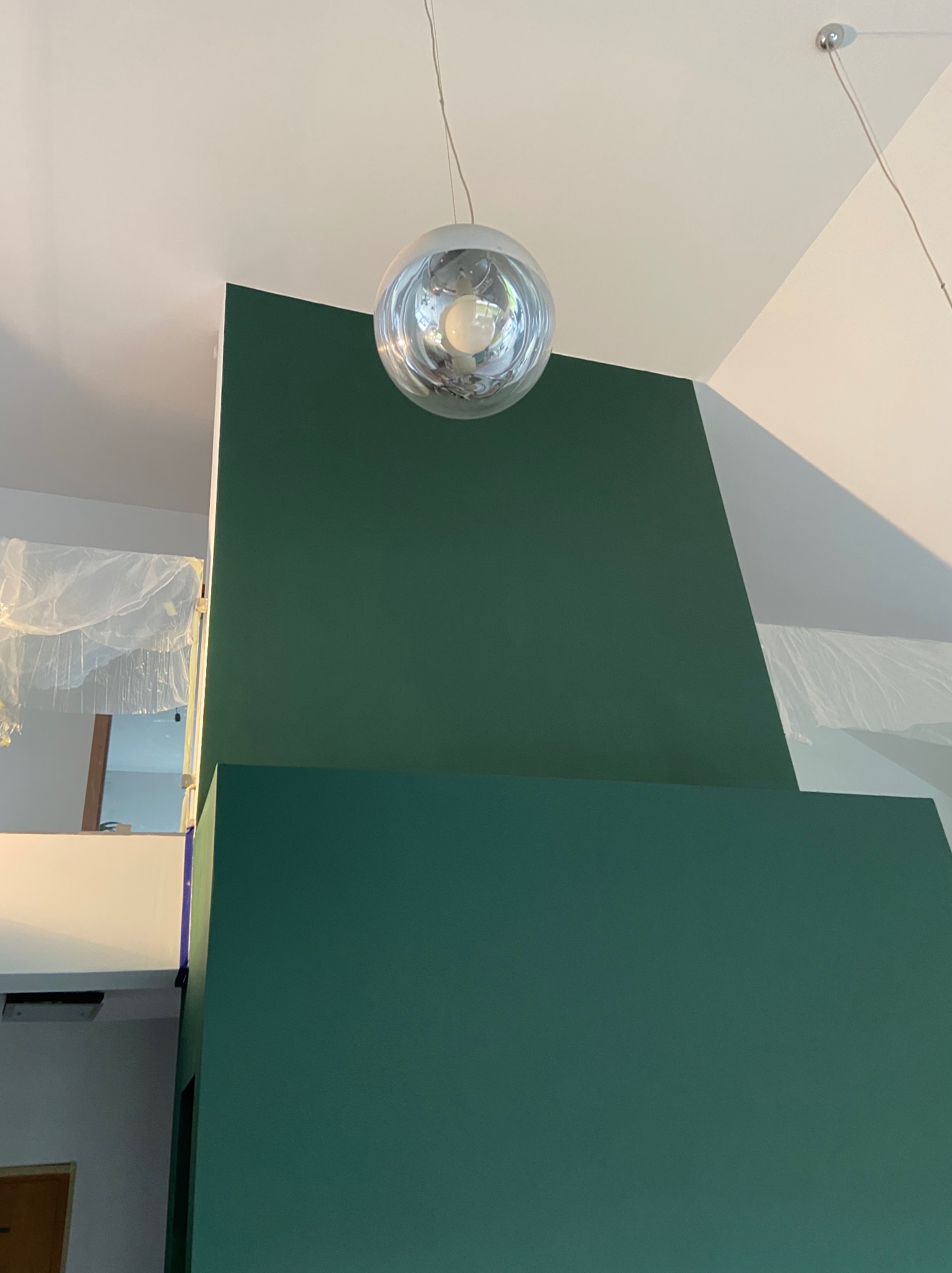 Pomieszczenie z fragmentem ściany pomalowanej na ciemnozielony kolor, widoczna lampa wisząca w kształcie kuli i zabezpieczona folią część pomieszczenia.