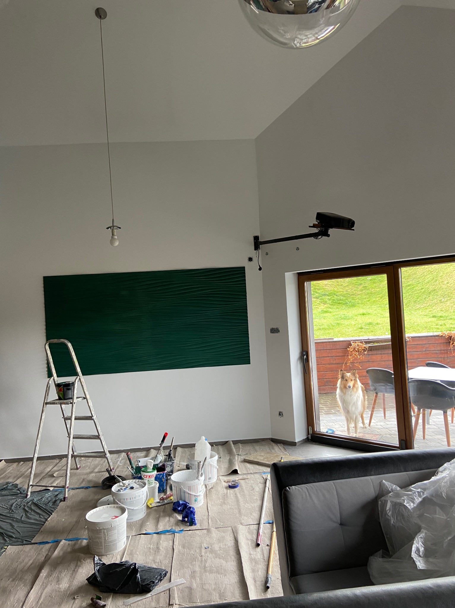 Wnętrze pokoju z zieloną, falistą ścianą w trakcie prac, drabina malarska, puszki z farbą, podłoga zabezpieczona kartonem, pies spoglądający przez okno tarasowe