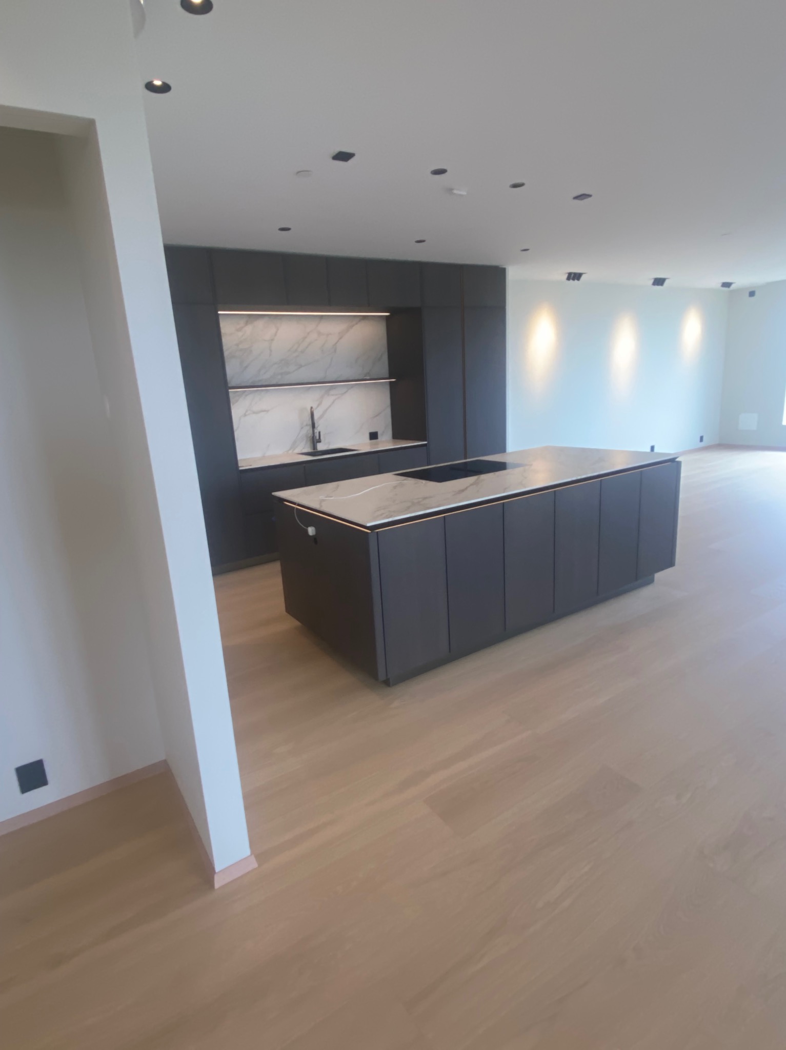 Nowoczesna, minimalistyczna kuchnia z wyspą i marmurowym blatem. Ciemne szafki, jasna podłoga, punktowe oświetlenie. Widok na przestronne, jasne wnętrze.