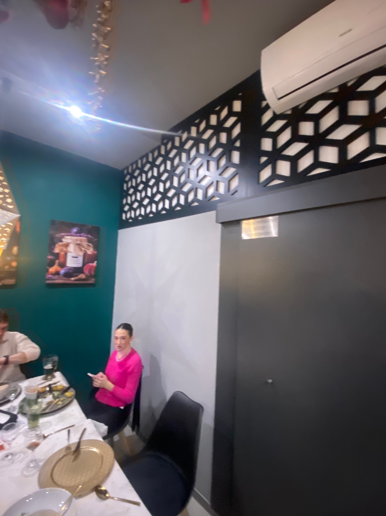 Nowoczesne wnętrze restauracji z dekoracyjną ścianą, klimatyzacją i obrazami. Widoczny stół z zastawą i siedzące osoby. Ściana w kolorze butelkowej zieleni.