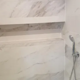 Wykończona kabina prysznicowa z półką wnękową, wyłożona płytkami imitującymi marmur, z chromowaną słuchawką prysznicową na ścianie.