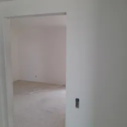 Wnętrze w trakcie remontu, widok przez futrynę drzwiową na pokój z białymi ścianami i przewodami na podłodze, gniazdko elektryczne bez obudowy na ścianie.