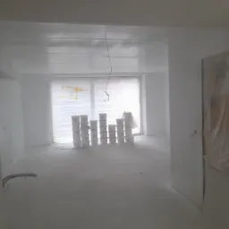 Wnętrze świeżo wykończonego pokoju, przygotowane do malowania, z oknem zabezpieczonym folią i ustawionymi rzędami puszek z farbą. Podłoga i ściany pokryte świeżą warstwą wykończeniową.