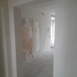 Wnętrze w trakcie remontu: świeżo pomalowane ściany na biało, zabezpieczone folią drzwi, widoczny kij oparty o ścianę w przejściu.