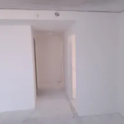 Wnętrze w trakcie prac wykończeniowych: białe ściany, surowy sufit z widocznymi przewodami, betonowa podłoga z resztkami zaprawy, widok na korytarz z otwartymi drzwiami.